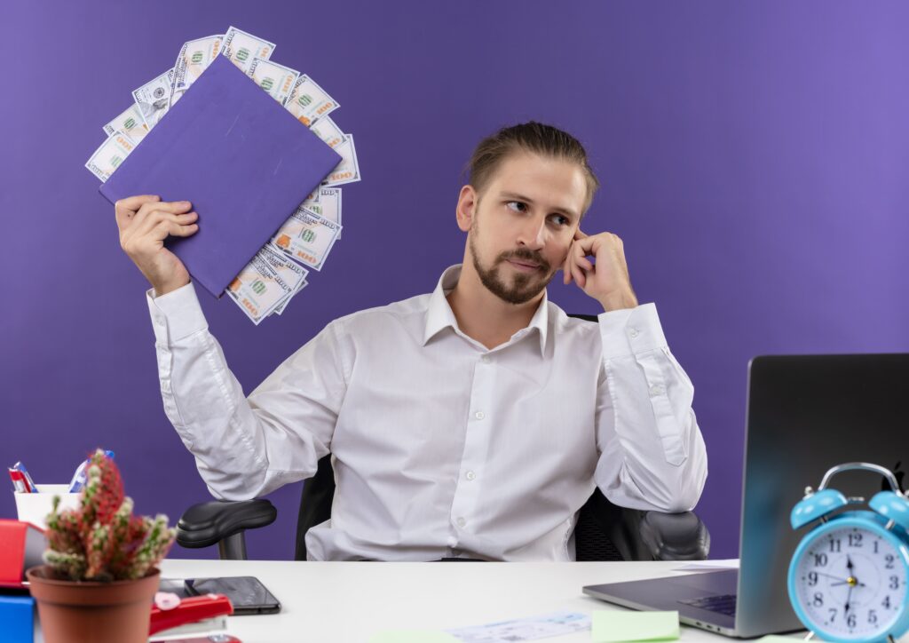 handsome businessman white shirt holding folder with cash looking aside with pensive expression sitting table offise purple background (1) min